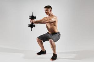 Confident fit man exercising with dumbbell while standing in squatting position against white background photo