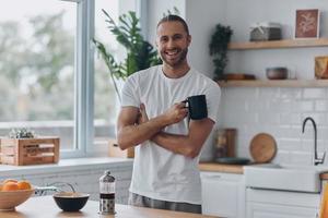 Handsome young man enjoying coffee and smiling while standing at the kitchen photo