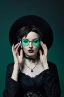 joven bruja de Halloween con un sombrero redondo y gafas redondas verdes sobre un fondo verde oscuro. cabello teñido, lápiz labial oscuro, aretes de luna creciente foto