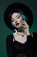 joven bruja de Halloween con un sombrero redondo y gafas redondas verdes sobre un fondo verde oscuro. cabello teñido, lápiz labial oscuro, aretes de luna creciente foto