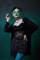 joven bruja de Halloween con un sombrero redondo y gafas redondas verdes sobre un fondo verde oscuro. cabello teñido, lápiz labial oscuro, aretes de luna creciente foto