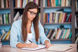 haciendo su tarea. mujer joven escribiendo algo en su bloc de notas y leyendo un libro mientras se sienta en el escritorio de la biblioteca foto