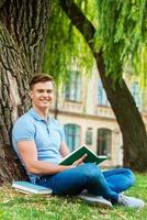 ratón de biblioteca confiado. estudiante varón confiado leyendo un libro y sonriendo mientras se sienta en el césped y frente al edificio de la universidad foto