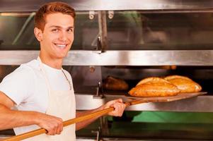 panadero confiado. joven confiado en delantal tomando pan recién horneado del horno y sonriendo foto