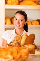solo la mejor y fresca panadería. Hermosa mujer joven en delantal sosteniendo una cesta con productos horneados mientras está de pie en la panadería. foto