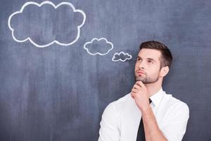 Flying of thoughts. Thoughtful young man holding hand on chin and looking away while standing against cloud chalk drawing on blackboard photo