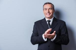 Celebrating success. Confident mature man in formalwear clapping hands and smiling while standing against grey background photo