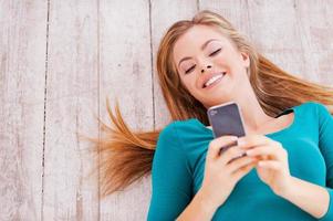 Lovely messages. Top view of beautiful young woman lying on the floor at her apartment holding phone photo