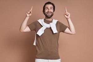 Handsome young man pointing up and smiling while standing against brown background photo