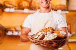 The freshest bread for you. Cropped image of young man in apron holding basket with baked goods and smiling while standing in bakery shop photo