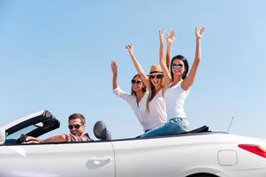 Spending great time together. Group of young happy people enjoying road trip in their white convertible while girls raising arms and smiling photo