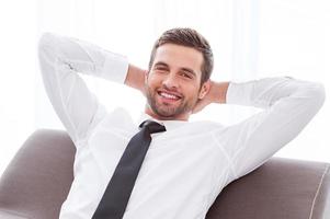 Taking time to relax. Happy young businessman in shirt and tie holding hands behind head and smiling while sitting at the chair photo