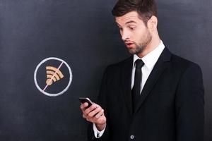 No Wi-Fi. Frustrated young man holding mobile phone and looking surprised because of bad connection while standing against sharing  symbol chalk drawing on blackboard photo