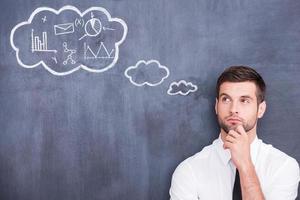 I need to think. Picture of handsome young man holding hand on chin and thinking while standing against cloud chalk drawing on blackboard photo