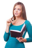 Looking for inspiration. Cheerful young women holding note pad and touching chin with pen while standing against white background photo