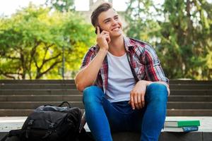 Good news from friend. Happy male student talking on the mobile phone and smiling while sitting at the outdoors staircase with books and backpack laying near him photo