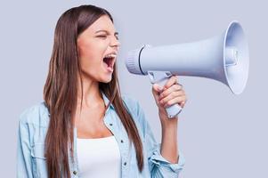 Want to be heard. Attractive young woman shouting through a megaphone, holding it in her hand while standing against grey background photo