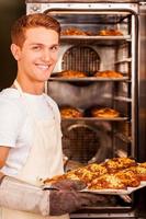 croissant recién horneado para ti. apuesto joven en delantal tomando los croissants recién horneados del horno y sonriendo foto