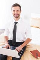 Working in hotel room. Top view of handsome young man in shirt and tie working on laptop and smiling while sitting on the bed in hotel room photo