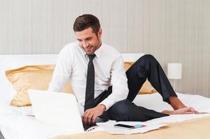 No time for rest. Handsome young man in shirt and tie working on laptop and smiling while sitting in bed at the hotel room photo