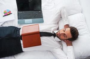 Businessman sleeping. Top view of handsome young man in shirt and tie holding hands behind head and keeping eyes closed while lying in bed at the hotel room photo