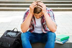 Failed again. Top view of frustrated male student touching his head with hands and looking down while sitting at the outdoors staircase with books and backpack laying near him photo