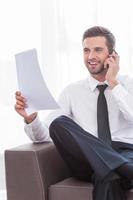 Discussing contract. Happy young businessman in shirt and tie talking on the mobile phone and holding document while sitting at the chair photo