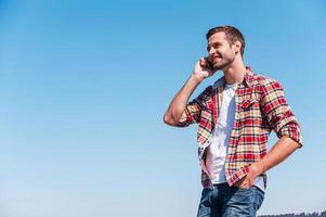 buena charla con un amigo. joven alegre hablando por teléfono móvil y sonriendo mientras está de pie al aire libre con el cielo azul como fondo foto