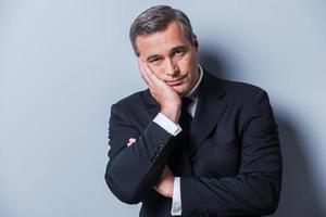 Bored businessman. Bored mature man in formalwear holding head in hand and looking at camera while standing against grey background photo