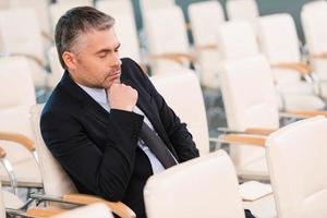 Boring conference. Bored mature man in formalwear holding hand on chin and keeping eyes closed while sitting on the chair in empty conference hall photo