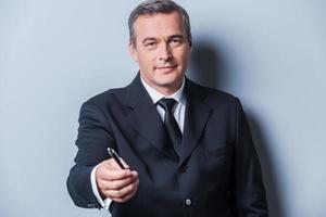 I need your signature. Portrait of confident mature man in formalwear stretching out a pen and smiling while standing against grey background photo