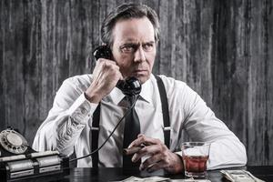 Tell me everything you know Serious senior man in shirt and suspenders talking on the old-fashioned telephone while sitting at the table and smoking cigar photo