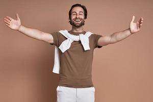 Cheerful young man keeping arms outstretched and smiling against brown background photo