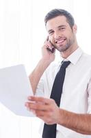 hombre de negocios confiado. vista de ángulo bajo de un joven alegre con camisa y corbata hablando por teléfono móvil y sosteniendo un documento foto