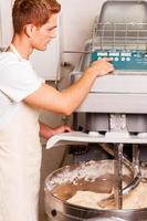 Working with dough. Confident young man in apron pushing button on dough mixing machine photo