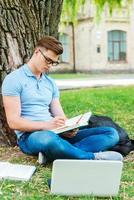 Spending every minute studying. Confident male student in glasses writing something in his note pad while sitting on the grass and in front of university building photo