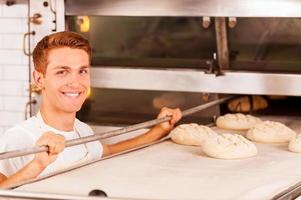 panadero confiado. confiado joven panadero poniendo masa en el horno y sonriendo foto