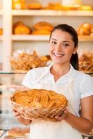 orgullosa de sus productos horneados. Hermosa mujer joven en delantal sosteniendo una cesta con productos horneados mientras está de pie en la panadería.