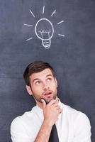 I got it. Thoughtful young man holding hand on chin and looking up while standing against blackboard with chalk drawing of light bulb photo