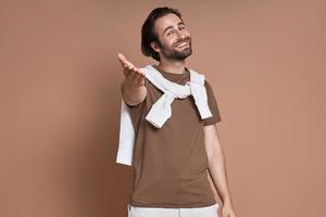 Cheerful young man keeping arm outstretched and smiling against brown background photo