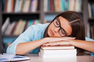 agotamiento del examen. mujeres jóvenes cansadas sosteniendo su cabeza en la pila de libros y durmiendo mientras están sentadas en el escritorio de la biblioteca foto