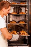 croissants recién horneados. confiado joven en delantal tomando los croissants recién horneados del horno foto