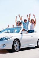 Friends and road trip. Group of young happy people enjoying road trip in their white convertible while raising arms and smiling photo