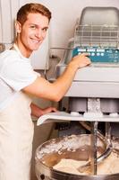 Preparing dough for pastry. Confident young man in apron pushing button on dough mixing machine and smiling photo