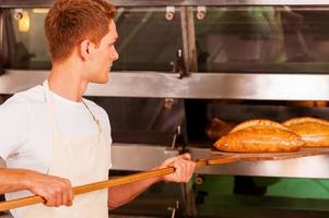 pan recién horneado. confiado joven en delantal tomando el pan recién horneado del horno foto