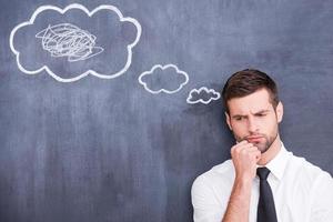 There is a mess in my head. Picture of confused handsome young man holding hand on chin and thinking while standing against cloud chalk drawing on blackboard photo