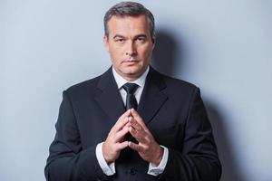 Confident business expert. Confident mature man in formalwear holding hands clasped and looking at camera while standing against grey background photo