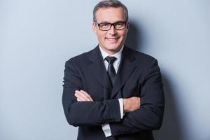 Confident and successful. Portrait of confident mature man in formalwear and eyeglasses looking at camera and smiling while standing against grey background photo