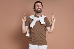 Handsome young man holding fingers crossed while standing against brown background photo