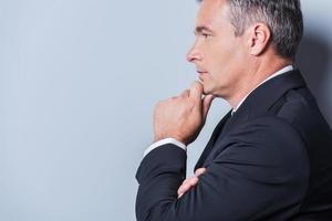 Lost in business thoughts. Side view of thoughtful mature man in formalwear holding hand on chin and looking away while standing against grey background photo